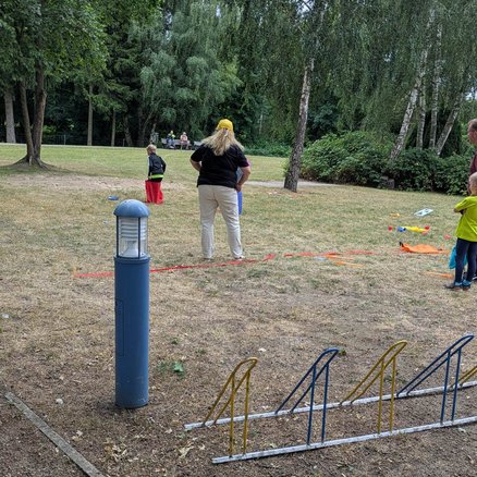 zeigt Kind beim Sackhüpfen auf einer Wiese und eine beobachtende Frau