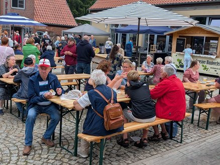 zeigt Markttreiben mit stehenden und an Holzgarnituren sitzenden Menschengruppen