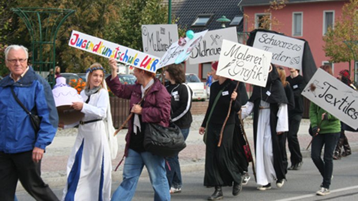 öffnet internen Link in aktuellem Fenster zeigt Ausschnitt des Festumzugs mit kostümierten Personen der Jugendkunstschule UER, die Schilder hochhalten