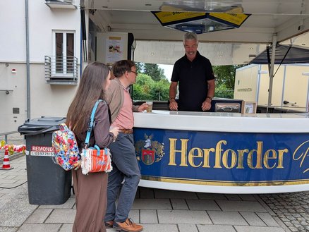 zeigt Getränkestand mit Person, die ausschenkt und Passanten