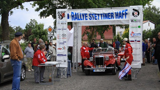 öffnet internen Link in aktuellem Fenster zeigt Starttor der Rallye mit einem abfahrbereiten Oldtimer und Rallyepersonal